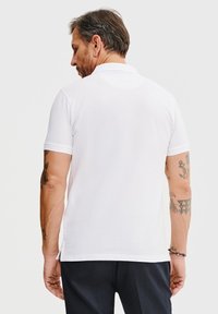 Biała koszulka polo z krótkim rękawem, z płaskim kołnierzykiem i bocznymi rozporkami. Na rękach widoczne tatuaże. Prosty, czysty design.
