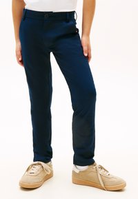 Bambino che indossa pantaloni aderenti blu navy, calzini bianchi e scarpe da ginnastica in suede beige, in piedi su uno sfondo chiaro uniforme.