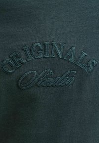 Zelena dukserica s izvezenim natpisima "ORIGINALS" i "Studio." Mekani materijal s glatkom površinom i klasičnim okruglim izrezom.