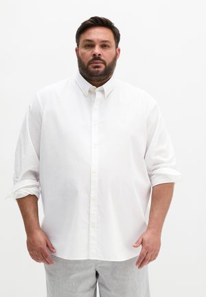 Brodaty mężczyzna o krótkich ciemnych włosach stojący, ubrany w białą koszulę na guziki z podwiniętymi rękawami i jasnoszare spodnie.