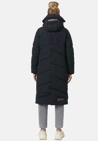 Long black puffer coat with a hood, featuring chevron quilting and an adjustable collar. Worn over gray jeans and white sneakers.
