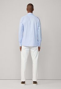 Camicia a righe blu chiaro con bottoni e maniche lunghe, pantaloni bianchi e sneakers casual; mostrata da dietro contro uno sfondo neutro.