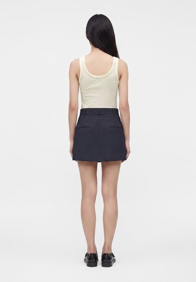 Femme aux longs cheveux foncés portant un haut beige sans manches, une mini-jupe bleu marine à fines rayures et des mocassins noirs, debout de dos.