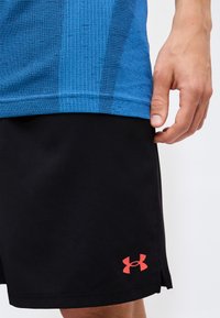 Χέρι που ακουμπάει κοντά σε μαύρα σορτς Under Armour με κόκκινο λογότυπο, φορώντας μπλε υφασμάτινη αθλητική μπλούζα, εμφανιζόμενο από τη μέση μέχρι το μέσο του μηρού.