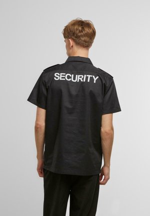 Melna īsām piedurknēm kreklā ar vārdu "SECURITY" lieliem baltiem burtiem uz muguras. Ar apkakles dizainu un plecu uzlikām.