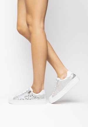 Jambes portant des baskets basses métalliques argentées avec des lacets blancs et des semelles blanches épaisses sur un fond clair uni.