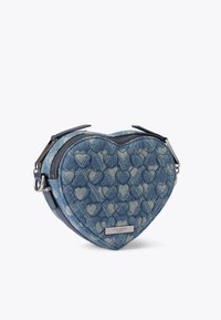 Sac en forme de cœur en denim avec texture matelassée, couleur bleue, deux fermetures éclair et accessoires en argent. Étiquette de la marque visible à l'avant.