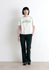 Cremefarvet cropped t-shirt med grøn "CALVIN KLEIN" logo, parret med sorte tætsiddende bukser og sorte sko. Minimalistisk design i neutrale nuancer.
