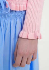 Ροζ πλεκτό πουλόβερ με ριμπ ύφανση και βολάν στα μανίκια, συνδυασμένο με φούστα σε ανοιχτό μπλε χρώμα, πλισέ, με σφηκωτή μέση και απαλό υφή.