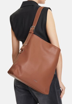 Žena v černém topu bez rukávů a tmavých džínách nesoucí velkou hnědou koženou kabelku přes rameno s nastavitelným popruhem.