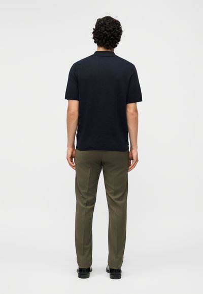 Άνδρας με μαλλιά με μπούκλες, ντυμένος με μαύρο κοντομάνικο πουκάμισο, ελαιώδες πράσινο παντελόνι και μαύρα παπούτσια, στέκεται με την πλάτη προς ένα λευκό φόντο.