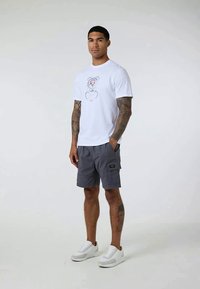 Ung man med tatuerade armar bär en vit grafisk t-shirt, grå cargobyxor och vita sneakers, står mot en enfärgad ljus bakgrund.
