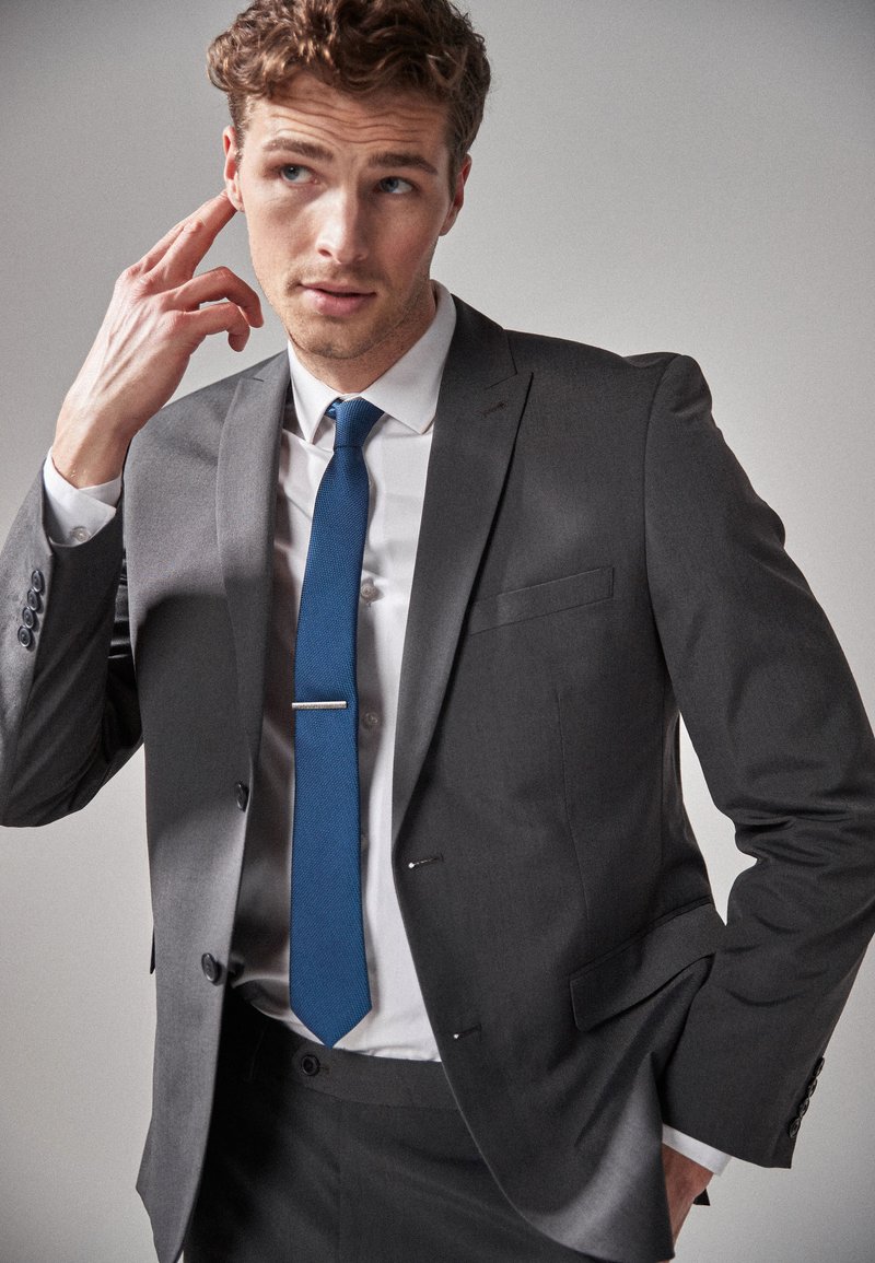 Costume gris ajusté, chemise blanche et cravate bleue texturée avec une pince à cravate en argent. Le modèle pose avec une main sur son visage.