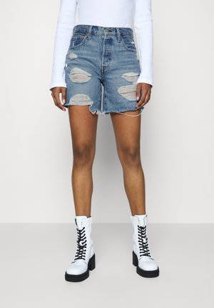 Person iført revet blå denimshorts, hvid langærmet top og hvide snørebåndsstøvler med sorte såler og snørebånd.