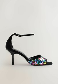 Sandal con tacco alto in suede nero con accentuazione di paillettes sulla parte anteriore. La scarpa presenta un cinturino alla caviglia regolabile e un design open toe.