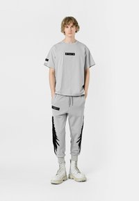 Camiseta de gran tamaño gris claro con etiqueta negra y pantalones de chándal a juego que presentan un estampado de plumas negras a lo largo de los lados, combinados con botas de combate blancas.