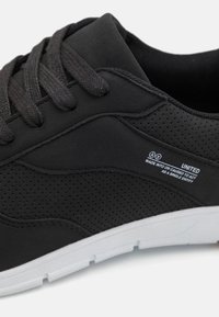 Black sneakers with textured upper, perforated design, and white rubber sole. Features black laces and branding label on the side.