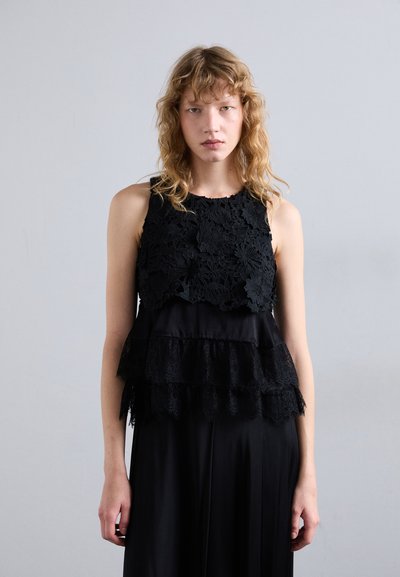 Top sans manches noir avec des détails en dentelle florale superposée sur la partie supérieure et un tissu lisse dans la partie inférieure.