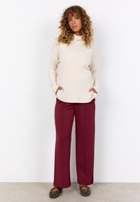 Maglione a costine color crema con colletto alto, abbinato a pantaloni a gamba larga bordeaux. L'outfit include tasche frontali e ballerine marroni con borchie.
