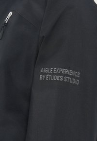 Mustmust kangas, millel on brodeeritud tekst "AIGLE EXPERIENCE BY ÉTUDES STUDIO" ning nähtav metallist tõmblukk vasakul ülal.