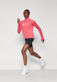 Ružičasta dugorukava sportska majica s visokim ovratnikom i bijelim logotipom, u kombinaciji s crnim šortsevima i bijelim tenisicama. Glatka, rastezljiva tkanina.