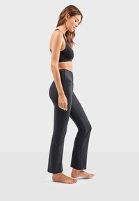 Zwarte, aansluitende yoga pants met een hoge tailleband, een gladde textuur en subtiele zijnaad, gedragen met een zwarte sportbeha, model in profielweergave.