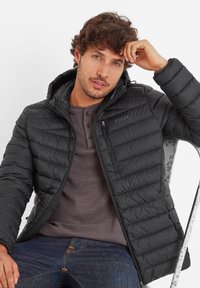 Black puffer jacket with a zippered pocket, quilted design, and adjustable hood, worn over a grey henley and dark jeans.