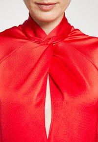 Top en satin rouge avec un détail en nœud au col et un décolleté en V profond, présentant une surface texturée qui reflète la lumière.