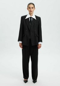 Črn pinstripes suknjič z dvojnim zapenjanjem in širokimi reverji, v kombinaciji s širokimi hlačami in belo srajco pod njim.
