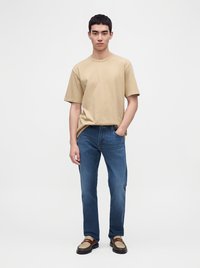 Jeune homme debout portant un t-shirt beige à manches courtes, un jean bleu et des mocassins marron bicolores, avec une main dans la poche, sur un fond uni.