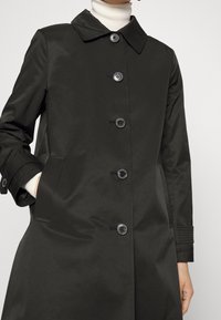 Svart knäppt kappa med krage, långa ärmar med knäppta manschetter och sidofickor, bärs över en vit polotröja.