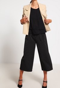 Blazer beige sopra un top nero a strati, abbinato a pantaloni a gamba larga neri con righe verticali. Tacchi neri eleganti completano il look.