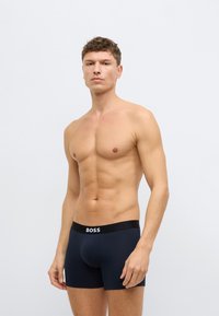 Σκούρα μπλε ανδρικά εσώρουχα κοντά με μαλακό ύφασμα, μαύρη επίπεδη ελαστική ζώνη μέσης με λευκό λογότυπο, εφαρμοστή σχεδίαση και λεία, ελαστική υφή.