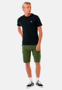 Maglietta nera a maniche corte con un logo verde, abbinata a pantaloni cargo verde oliva. Calzini bianchi e sneakers nere completano l'outfit.