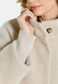 Beige, textuurjasje met grote knoopdetails en ribbelgebreide manchetten. De stof lijkt zacht en harig, wat warmte en comfort biedt.