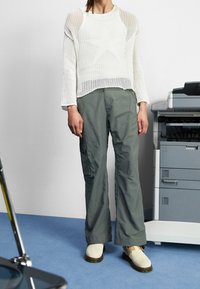 Suéter blanco de punto con un patrón de estrellas; pantalones cargo verde oliva; zapatos de tela beige. Con un fondo de oficina minimalista.