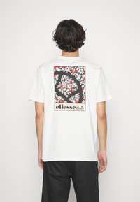 Weißes Baumwoll-T-Shirt mit einem bunten Blumenmuster auf der Rückseite, einem schwarzen Hintergrund und dem "ellesse"-Logo am unteren Ende.