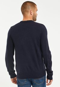 Tmavě modrý pletený svetr s dlouhými rukávy, kulatým výstřihem a vroubkovanými manžetami. Hladká textura; nošený s džíny, s pohledem zezadu.