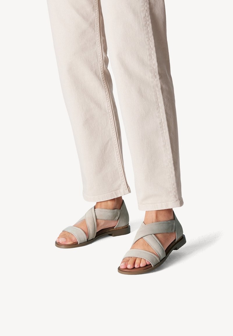 Pieds portant des sandales beige clair à brides croisées associées à un pantalon droit écru sur un fond uni.