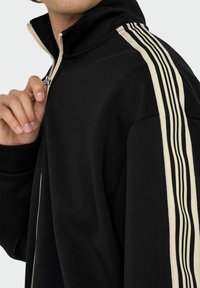Schwarze Zip-Jacke aus weichem Stoff. Verfügt über einen hohen Kragen und kontrastierende cremefarbene vertikale Streifen an den Ärmeln; sportliches Design mit einer taillierten Form.