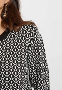 Pull en noir et blanc à motifs avec un motif en losange, col rond et poignets côtelés. Le tissu semble texturé et tricoté.