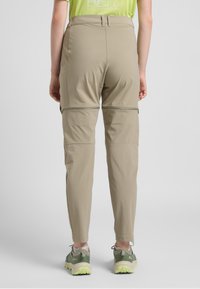 Pantaloni da escursionismo beige con cerniera, dal taglio affusolato, caratterizzati da una texture liscia e dettagli delle cuciture in vista. Indossati con scarpe da escursionismo grigio-verdi.