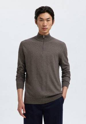 Homme portant un pull marron à demi-fermeture éclair et un pantalon foncé, debout avec une main dans la poche devant un fond clair uni.