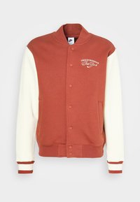 Nike varsityjacka med rostfärgad överdel och krämfärgade ärmar; har ribbade muddar, två framfickor och en broderad logotyp på bröstet.