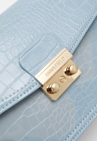 Světle modrá psaníčko s krokodýlovou texturou a zlatým zapínáním, na kterém je uveden název značky. Hladký materiál a detaily kontrastního prošití.