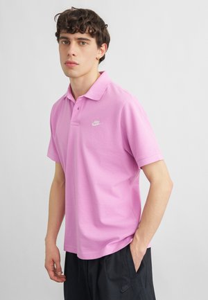 Młody mężczyzna ubrany w jasnofioletową koszulkę polo Nike i czarne spodnie Nike, stoi z jedną ręką w kieszeni na jednolitym tle.