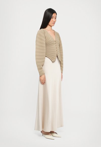 Beige cardigan with pleated design, V-neck, and buttons. Paired with a long, flowing cream silk skirt and pale pointed-toe shoes.