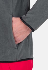 Felpa in pile grigia con tasca laterale, dotata di superficie testurizzata e bordi neri sui polsini e sull'orlo, indossata con pantaloni rossi.