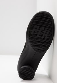 Chaussure en cuir noir présentant un talon arrondi et une semelle en caoutchouc texturée avec des motifs circulaires. Marque visible sur la semelle.