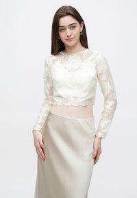 Valkoinen pitsinen lyhyt paita, jossa kukkakuosi ja pitkät hihat, yhdistetty satiiniseen beigeen hameeseen. Reunassa on aaltoileva kuvio ja pinta on sileä.